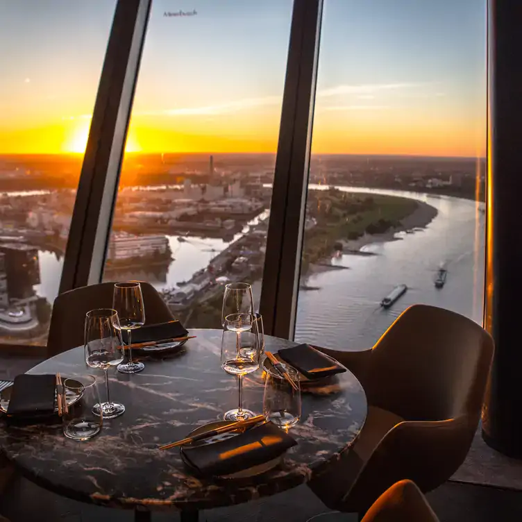 Tisch mit Gedecken und Weingläsern vor dem atemberaubenden Ausblick auf den Sonnenuntergang und den Rhein vom Qomo Restaurant und Bar im Fernsehturm, einem der besten Restaurants in Düsseldorf.