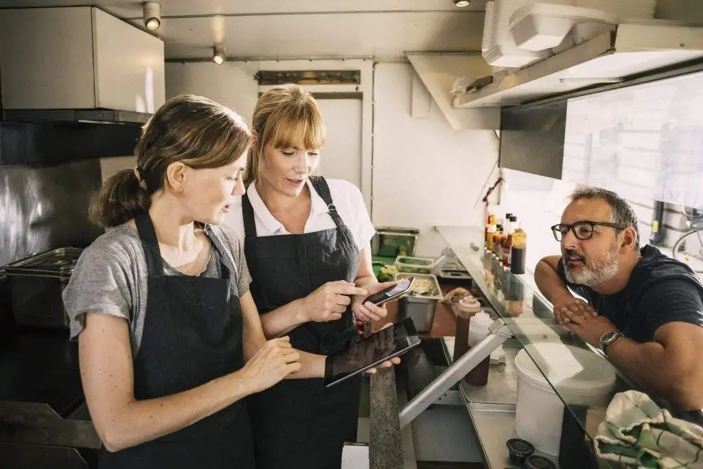 Das Bild zeigt zwei Angestellte im Restaurant, die hinter einer Theke stehen. Eine der Angestellten trägt ein weißes Poloshirt und eine schwarze Schürze und zeigt der Kollegin etwas auf einem Telefon. Die Kollegin trägt ein graues T-Shirt und eine schwarze Schürze und hält ein Tablet in der Hand. Ein Kunde mit einer schwarz umrandeten Brille lehnt sich über den Tresen.